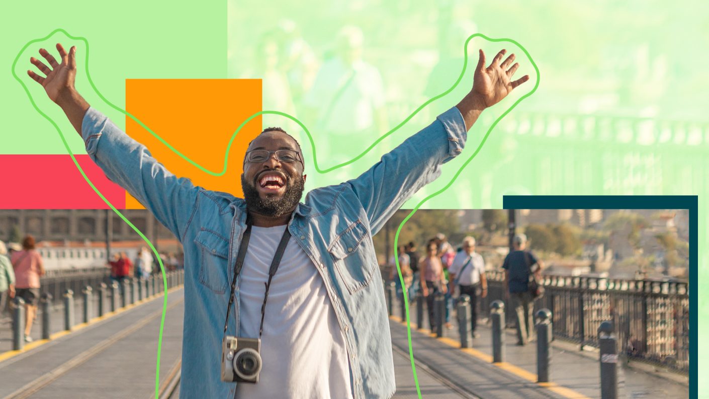 Man standing on a bridge, smiling with his arms in the air, carrying a camera around his neck