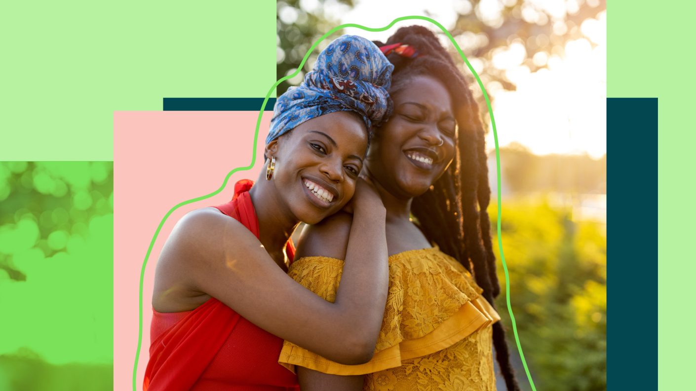 Two people with colourful clothes both smiling, one leaning on the back of the other one.