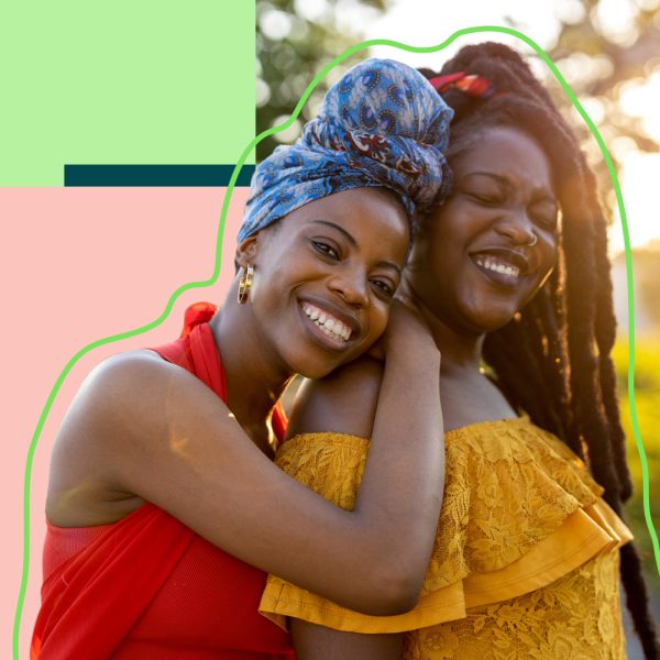 Two people with colourful clothes both smiling, one leaning on the back of the other one.