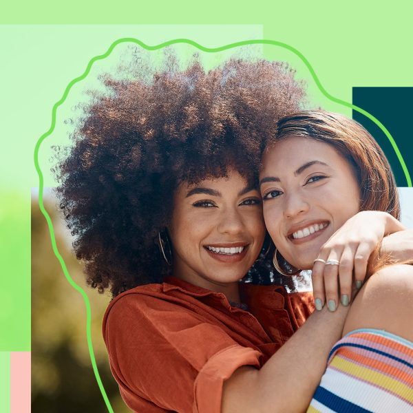 Two women smiling and hugging. The left woman has afro hair and wears an orange shirt. The right woman has Asian roots and wears a striped off-shoulder top.